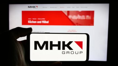 Stuttgart, Almanya - 07-31-2024: Alman hizmet şirketi MHK Group AG 'nin logosuyla cep telefonu tutan kişi iş sayfasının önünde. Telefon ekranına odaklan.