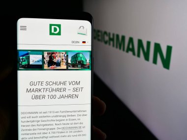 Stuttgart, Almanya - 08-01-2024: Alman ayakkabı perakende şirketi Deichmann SE 'nin internet sayfası ile logo önünde cep telefonu tutan kişi. Telefon ekranının merkezine odaklan.