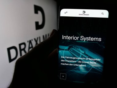 Stuttgart, Germany - 08-02-2024: Person holding mobile phone with logo of German automotive company Fritz Draexlmaier GmbH Co. KG in front of web page. Focus on phone display. clipart