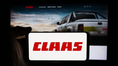 Stuttgart, Germany - 08-02-2024: Person holding cellphone with logo of German agricultural machinery company Claas KGaA mbH in front of business webpage. Focus on phone display. clipart