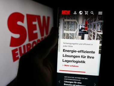 Stuttgart, Germany - 08-05-2024: Person holding cellphone with webpage of German engineering company SEW-Eurodrive GmbH Co. KG in front of logo. Focus on center of phone display. clipart