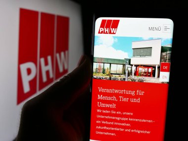 Stuttgart, Germany - 08-05-2024: Person holding cellphone with webpage of German meat processing company Lohmann Co. AG (PHW-Gruppe) with logo. Focus on center of phone display. clipart