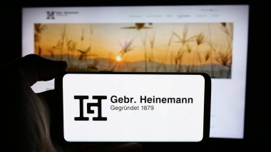 Stuttgart, Almanya - 08-06-2024: Alman seyahat perakende şirketi Gebr 'in logosuna sahip cep telefonu sahibi kişi. Heinemann SE Co. KG web sayfasının önünde. Telefon ekranına odaklan.