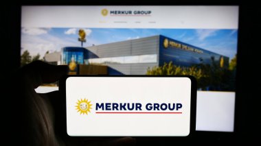 Stuttgart, Almanya - 08-06-2024: Alman eğlence şirketi Merkur Group 'un logosuyla cep telefonu tutan kişi iş sayfasının önünde. Telefon ekranına odaklan.