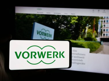 Stuttgart, Germany - 08-07-2024: Person holding mobile phone with logo of German household appliances company Vorwerk SE Co. KG in front of web page. Focus on phone display. clipart