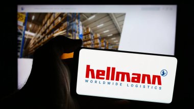 Stuttgart, Germany - 08-07-2024: Person holding mobile phone with logo of German company Hellmann Worldwide Logistics SE Co. KG in front of web page. Focus on phone display. clipart