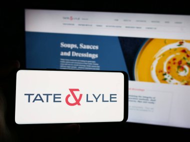 Stuttgart, Germany - 08-16-2024: Person holding smartphone with logo of British food processing company Tate and Lyle plc in front of website. Focus on phone display. clipart