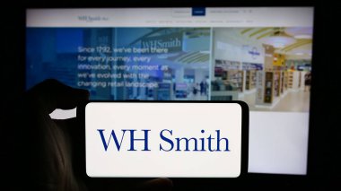 Stuttgart, Germany - 08-16-2024: Person holding mobile phone with logo of British retail company WH Smith plc (WHSmith) in front of business web page. Focus on phone display. clipart