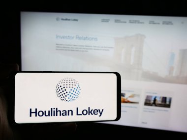 Stuttgart, Germany - 08-16-2024: Person holding mobile phone with logo of American investment banking company Houlihan Lokey Inc. in front of web page. Focus on phone display. clipart