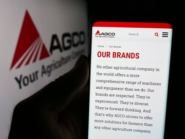 Stuttgart, Almanya - 08-18-2024: ABD tarım makineleri şirketi AGCO Corporation 'ın web sayfası ile logo önünde cep telefonu tutan kişi. Telefon ekranının merkezine odaklan.