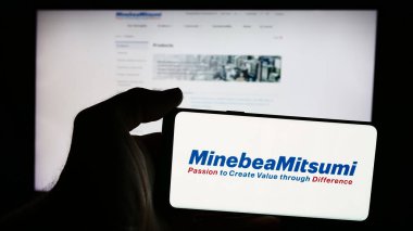 Stuttgart, Germany - 08-21-2024: Person holding mobile phone with logo of Japanese electronics company MinebeaMitsumi Inc. in front of business web page. Focus on phone display. clipart