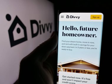 Stuttgart, Germany - 08-26-2024: Person holding cellphone with webpage of US real estate company Divvy Homes Inc. in front of business logo. Focus on center of phone display. clipart