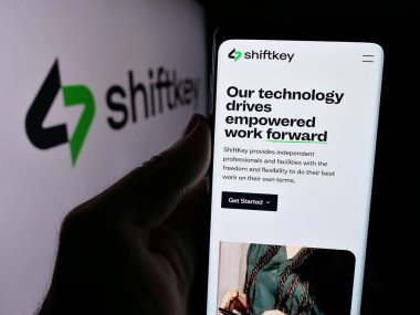 Stuttgart, Germany - 08-27-2024: Person holding cellphone with webpage of US healthcare platform company ShiftKey LLC in front of business logo. Focus on center of phone display. clipart