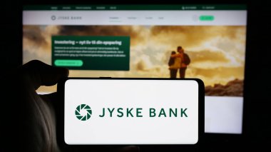 Stuttgart, Germany - 09-27-2024: Person holding mobile phone with logo of Danish financial company Jyske Bank AS on screen in front of business web page. Focus on phone display. clipart