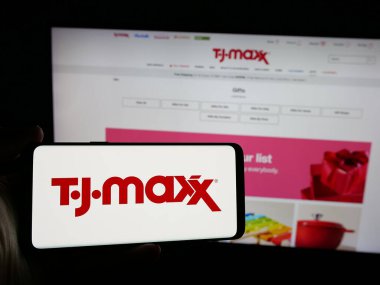 Stuttgart, Germany - 10-04-2024: Person holding mobile phone with logo of US discount department store company TJ Maxx on screen in front of web page. Focus on phone display. clipart