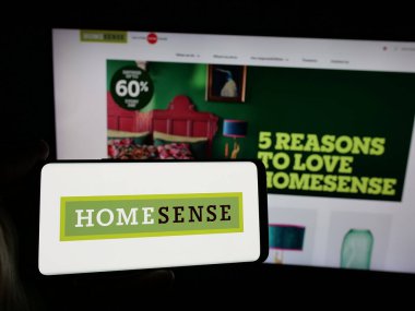 Stuttgart, Germany - 10-04-2024: Person holding smartphone with logo of furnishing store company HomeSense on screen in front of website. Focus on phone display. clipart