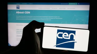 Stuttgart, Germany - 10-07-2024: Person holding mobile phone with logo of European Committee for Standardization (CEN) on screen in front of web page. Focus on phone display. clipart