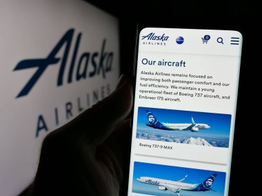 Stuttgart, Germany - 10-08-2024: Person holding smartphone with webpage of US airline company Alaska Air Group Inc. on screen in front of logo. Focus on center of phone display. clipart