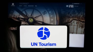 Stuttgart, Germany - 10-09-2024: Person holding cellphone with logo of United Nations agency UN Tourism on screen in front of webpage. Focus on phone display. clipart