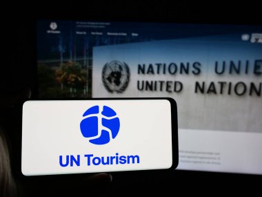 Stuttgart, Germany - 10-09-2024: Person holding smartphone with logo of United Nations agency UN Tourism on screen in front of website. Focus on phone display. clipart