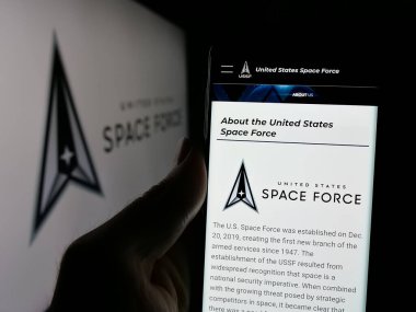 Stuttgart, Germany - 2024-10-10: Person holding smartphone with webpage of United States Space Force (USSF) on screen in front of logo. Focus on center of phone display. clipart