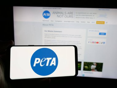 Stuttgart, Germany - 10-11-2024: Person holding smartphone with logo of People for the Ethical Treatment of Animals (PETA) on screen in front of website. Focus on phone display. clipart