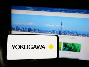 Stuttgart, Germany - 10-14-2024: Person holding mobile phone with logo of Japanese company Yokogawa Electric Corporation on screen in front of web page. Focus on phone display. clipart