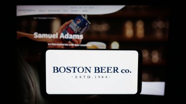 Stuttgart, Germany - 10-17-2024: Person holding cellphone with logo of US brewery business Boston Beer Company on screen in front of business webpage. Focus on phone display. clipart