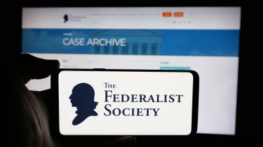 Stuttgart, Germany - 10-28-2024: Person holding cellphone with logo of US legal organisation Federalist Society on screen in front of webpage. Focus on phone display. clipart