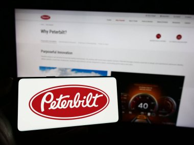 Stuttgart, Almanya - 12-27-2023: İnternet sayfası önünde üzerinde iş yapmayan Peterbilt Motor Şirketi 'nin logosu olan cep telefonu tutan kişi. Telefon ekranına odaklan.