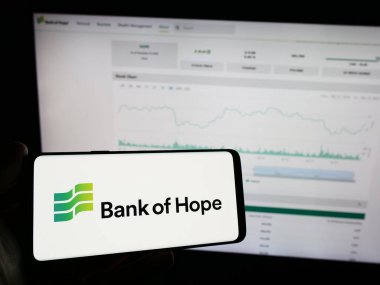 Stuttgart, Germany - 12-23-2023: Person holding mobile phone with logo of American financial company Hope Bancorp Inc. (Bank of Hope) in front of web page. Focus on phone display. clipart