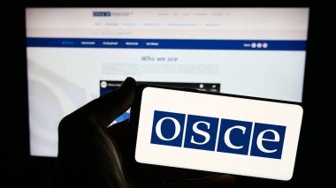 Stuttgart, Germany - 12-28-2023: Person holding smartphone with logo of Organization for Security and Co-operation in Europe (OSCE) in front of website. Focus on phone display. clipart