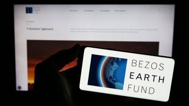 Stuttgart, Germany - 12-29-2023: Person holding mobile phone with logo of environmental organization Bezos Earth Fund in front of web page. Focus on phone display. clipart