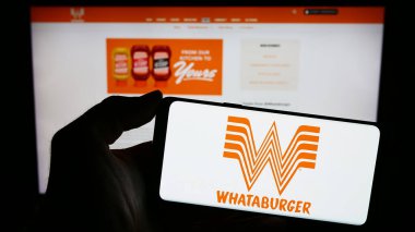 Stuttgart, Germany - 12-31-2023: Person holding mobile phone with logo of American fast food company Whataburger Restaurants LLC in front of web page. Focus on phone display. clipart