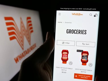 Stuttgart, Germany - 12-31-2023: Person holding cellphone with webpage of US fast food company Whataburger Restaurants LLC in front of logo. Focus on center of phone display. clipart