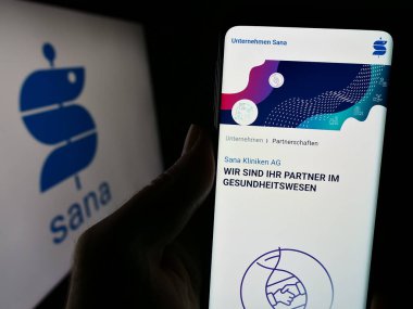 Stuttgart, Almanya - 12-31-2023: Alman sağlık şirketi Sana Kliniken AG 'nin internet sayfası ile iş logosu önünde cep telefonu tutan kişi. Telefon ekranının merkezine odaklan.