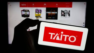 Stuttgart, Almanya - 02-03-2024: Japon video oyunları şirketi Taito Corporation 'ın logosuyla cep telefonu tutan kişi iş sayfasının önünde. Telefon ekranına odaklan.