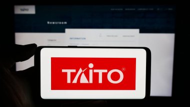 Stuttgart, Almanya - 02-03-2024: Japon video oyunları şirketi Taito Corporation 'ın logosunun bulunduğu cep telefonu sahibi kişi iş sayfasının önünde. Telefon ekranına odaklan.