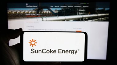 Stuttgart, Germany - 02-11-2024: Person holding mobile phone with logo of American coke producing company SunCoke Energy Inc. in front of web page. Focus on phone display. clipart