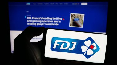 Stuttgart, Germany - 02-13-2024: Person holding smartphone with logo of French lottery company Francaise des Jeux (FDJ) in front of website. Focus on phone display. clipart