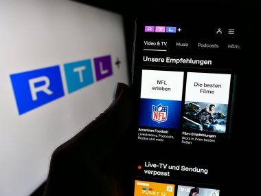 Stuttgart, Germany - 02-13-2024: Person holding cellphone with webpage of German entertainment platform RTL plus in front of business logo. Focus on center of phone display. clipart