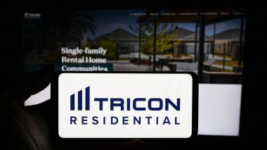 Stuttgart, Germany - 02-13-2024: Person holding mobile phone with logo of Canadian real estate company Tricon Residential Inc. in front of web page. Focus on phone display. clipart