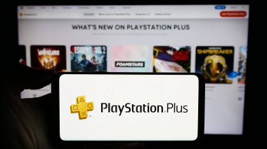 Stuttgart, Germany - 02-14-2024: Person holding cellphone with logo of subscription service PlayStation Plus (PS Plus) in front of business webpage. Focus on phone display. clipart