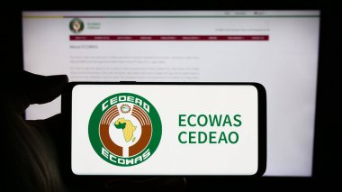 Stuttgart, Germany - 02-15-2024: Person holding cellphone with logo of Economic Community of West African States (ECOWAS) in front of webpage. Focus on phone display. clipart