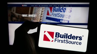 Stuttgart, Almanya - 02-22-2024: Web sayfası önünde üzerinde Amerikan inşaat malzemeleri şirketi Builders First Source Inc. 'in logosu olan cep telefonu sahibi kişi. Telefon ekranına odaklan.