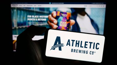 Stuttgart, Almanya - 02-22-2024: Amerikan içecek şirketi Athletic Brewing Company 'nin logosunun bulunduğu cep telefonu sahibi kişi web sayfasının önünde. Telefon ekranına odaklan.