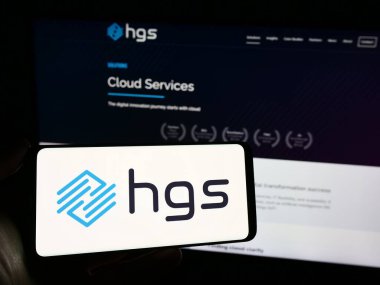 Stuttgart, Almanya - 02-24-2024: Hint Hinduja Global Solutions Limited (HGS) şirketinin logosuna sahip cep telefonu sahibi kişi web sayfası önünde. Telefon ekranına odaklan.