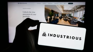 Stuttgart, Almanya - 02-26-2024: İş dünyası web sayfasının önünde üzerinde çalışan Amerikan iş birliği şirketi Industrious 'un logosu olan mobil pofon tutan kişi. Telefon ekranına odaklan.