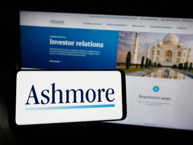 Stuttgart, Germany - 03-16-2024: Person holding mobile phone with logo of British investment management company Ashmore Group plc in front of web page. Focus on phone display. clipart