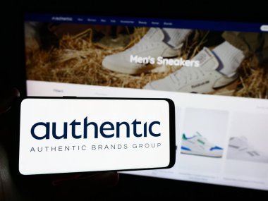 Stuttgart, Almanya - 03-17-2024: Amerikan şirketi Authentic Brand Group LLLC 'nin (ABG) logosuna sahip cep telefonu sahibi kişi iş sayfasının önünde. Telefon ekranına odaklan.